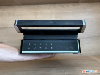 Een persoon houdt een strakke, zwarte luchtreiniger met een modern design vast. De luchtreiniger is voorzien van een digitaal bedieningspaneel met aanraakgevoelige knoppen en pictogrammen. De achtergrond is een licht houten oppervlak met rechtsonder het Keurio.nl-logo zichtbaar.