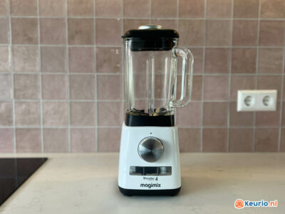 Een Magimix Blender 4 staat op een aanrecht in een moderne keuken met een roze betegelde achterwand. De blender heeft een transparante glazen kan, een zwart deksel en een witte basis met een grote zilveren wijzerplaat. Op de muur erachter is een stopcontact zichtbaar.