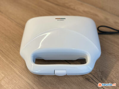 Een witte Domo tosti-ijzer geplaatst op een houten oppervlak. Het apparaat heeft een gesloten deksel met een handvat en een indicatielampje aan de bovenzijde. Aan de achterkant steekt een zwart netsnoer uit. Rechtsonder in de afbeelding staat ook een klein logo van keurio.nl.