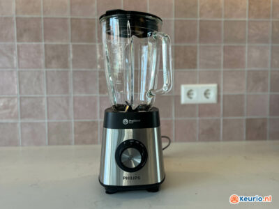 Op een aanrecht staat een zilverkleurige en zwarte Philips-blender met een betegelde achterwand op de achtergrond. De blender heeft een glazen kan en een grote draaiknop op de basis met meerdere instellingen. Op de achterwand bevindt zich een stopcontact. Het logo van Keurio.nl staat rechtsonder.