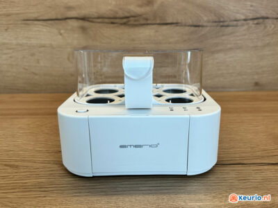 Een witte Emerio eierkoker wordt op een houten oppervlak geplaatst. Het apparaat heeft een transparant deksel, sleuven voor maximaal zes eieren en een handvat aan de bovenkant. Op de voorkant van het fornuis staat een logo en dit geeft aan dat het van keurio.nl is.
