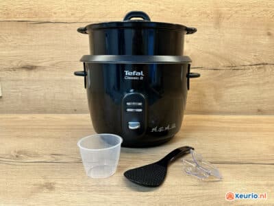 Een Tefal Classic 2 rijstkoker wordt op een houten oppervlak geplaatst met een kleine transparante maatbeker, een zwarte rijstschep en een plastic lepelsteun ervoor. De achtergrond is voorzien van een gestructureerd houten paneel.