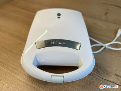 Een witte Tristar tosti-ijzer staat op een houten ondergrond. Het apparaat is voorzien van een metalen strip met de merknaam, een aan/uit-indicatielampje en een antisliphandgreep. Er is een netsnoer zichtbaar naast het apparaat.