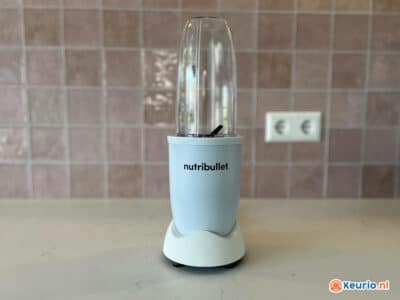 Een NutriBullet-blender met een strakke witte basis en een doorzichtige plastic mengbeker wordt op een lichtgekleurd aanrecht geplaatst. De achtergrond is voorzien van een roze betegelde muur en een wit stopcontact.