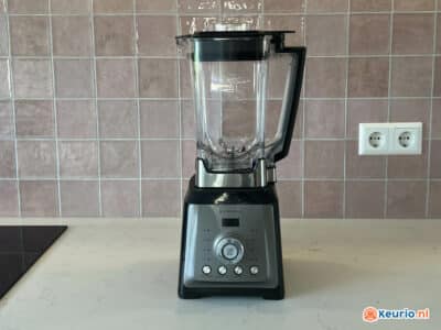 Een zwart-zilveren blender staat op een lichtgekleurd aanrecht tegen een roze betegelde achterwand. De blender heeft een doorzichtige container met verschillende bedieningsknoppen op de basis. Aan de linkerkant is gedeeltelijk een kookplaat zichtbaar, samen met een dubbel stopcontact aan de muur.