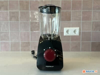 Een zwarte Aigostar-blender met een rode wijzerplaat staat op een wit aanrecht voor een betegelde achterwand. De blender heeft een doorzichtige glazen kan en een netsnoer dat aan de achterkant uitsteekt. De achtergrond is voorzien van een stopcontact en lichtroze tegels.