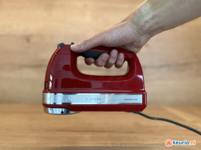 Een hand die een rode elektrische handmixer met 9 snelheden vasthoudt tegen een houten achtergrond. De mixer heeft een glanzende metalen strip en is op de zijkant voorzien van het opschrift "Assembled in USA". Rechtsonder in de afbeelding is het logo "keurio.nl" zichtbaar.