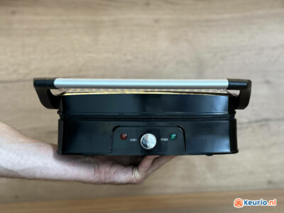 Een hand houdt een zwarte elektrische grill tegen een houten oppervlak. De grill heeft een zilveren handvat aan de bovenkant, een centrale draaiknop voor temperatuurregeling met het opschrift "min" en "max", en twee indicatielampjes, een rood en een groen.