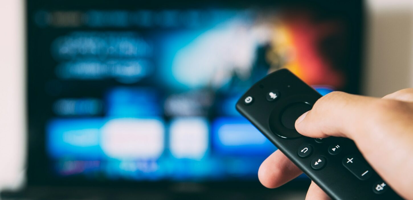Een close-up van een hand die een afstandsbediening vasthoudt, wijst naar een televisie met een kleurrijk streamingservicemenu, geoptimaliseerd met SEO-trefwoorden.