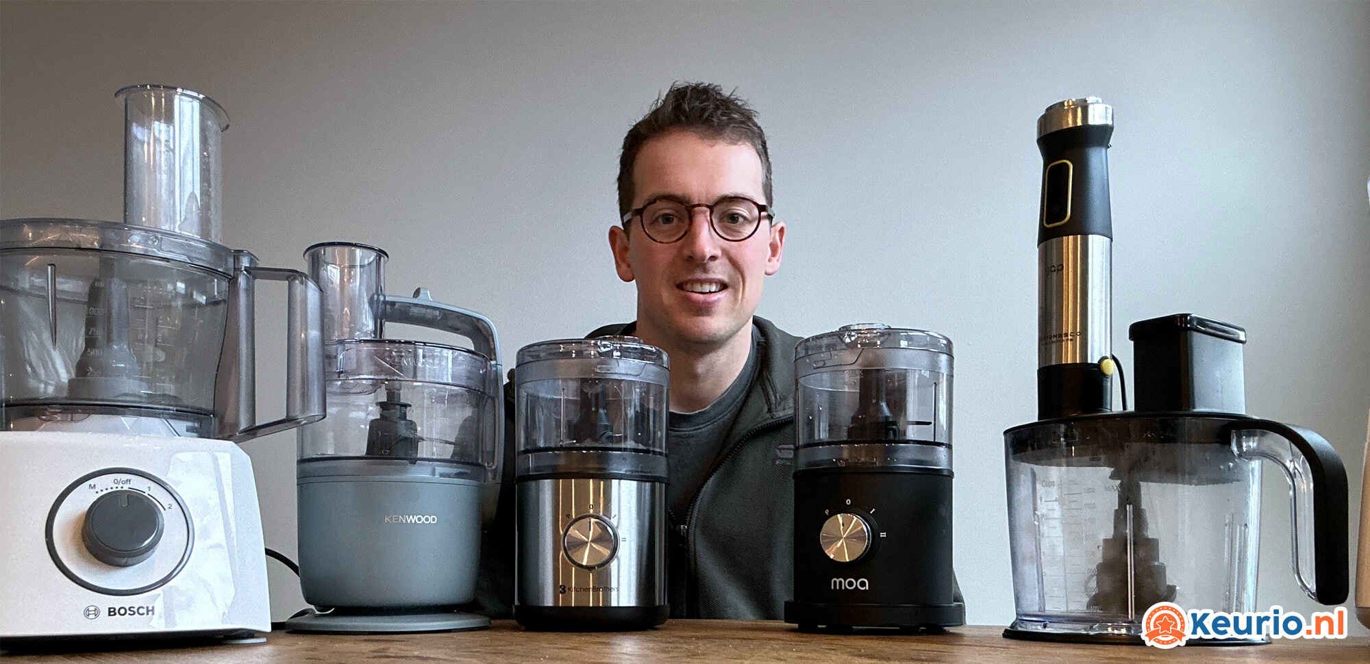 Een jonge man met een bril glimlacht naar de camera, zittend achter een tafel met een verzameling verschillende keukenapparatuur, waaronder meerdere keukenmachines en blenders.