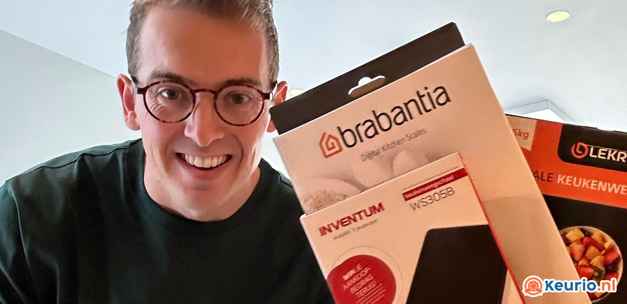 Een glimlachende man met een bril die twee dozen met keukenproducten omhoog houdt, waaronder keukenweegschalen, de ene met het opschrift 'brabantia' en de andere met 'inventum', met een logo zichtbaar voor keurio.nl in een huiselijke setting.