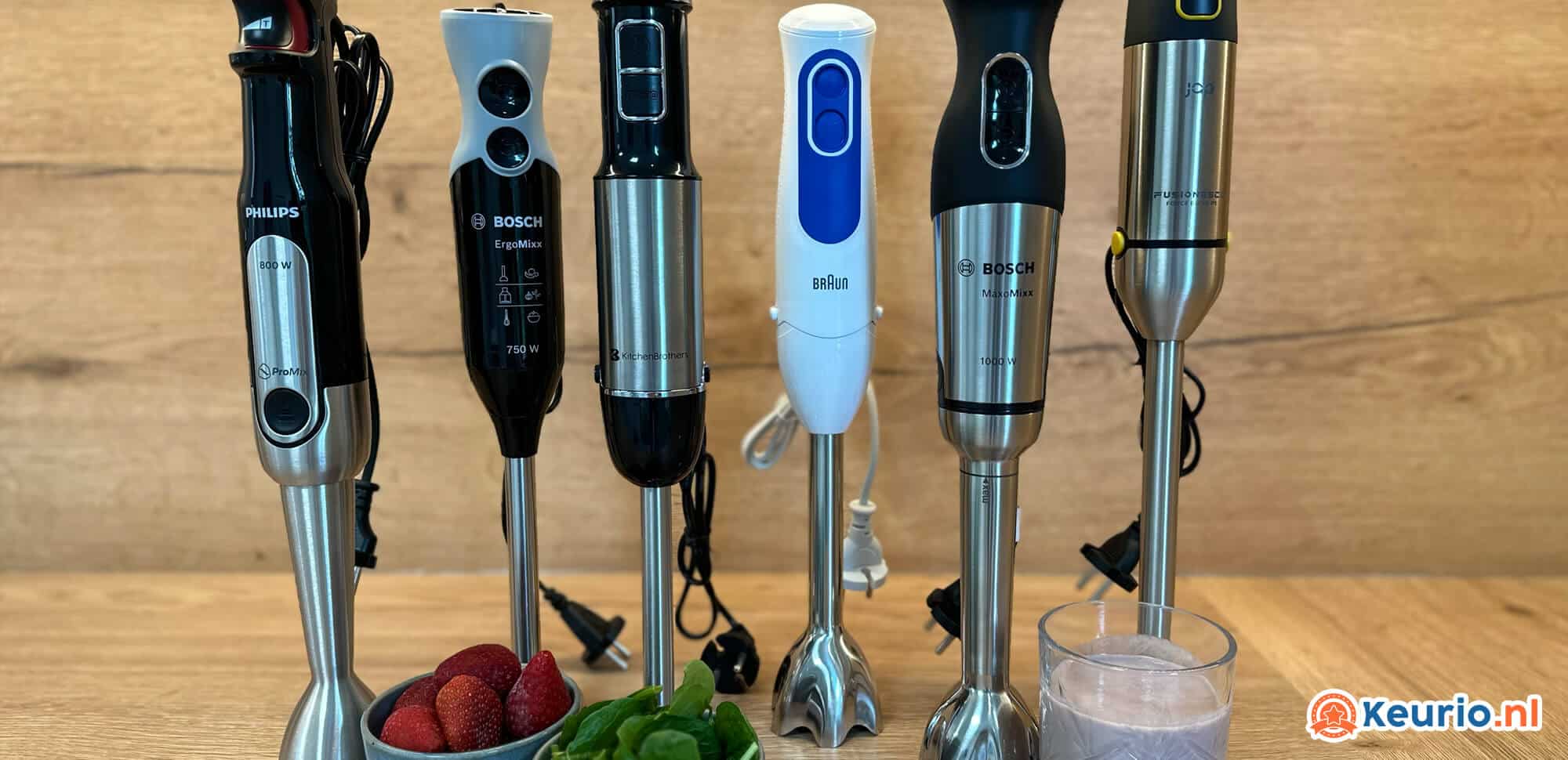 Op een houten aanrecht staat een opstelling van zes verschillende staafmixers van merken als Philips, Bosch en Braun. Op de toonbank staat ook een glas smoothie, een schaal met aardbeien en een bosje spinazie. Deze indrukwekkende collectie keukenapparaten valt op tegen de rustieke houten achtergrond.