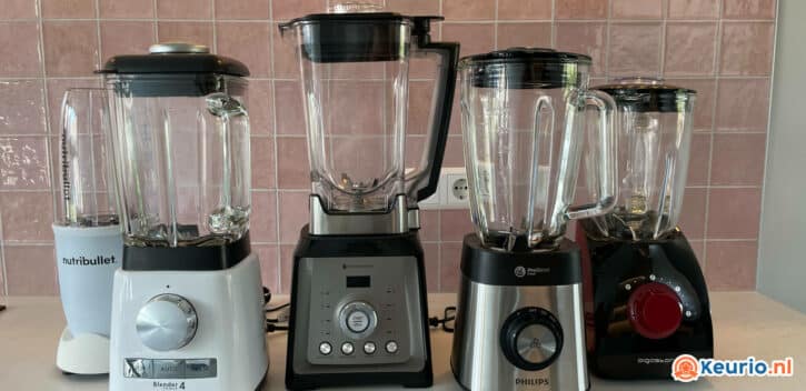Een opstelling van vijf verschillende aanrechtblenders wordt tentoongesteld tegen een roze betegelde muur. Van links naar rechts zijn de merken NutriBullet, Silvercrest, ProfiCook, Philips en AEG. Elke blender is voorzien van een doorzichtige glazen kan en verschillende bedieningsknoppen.