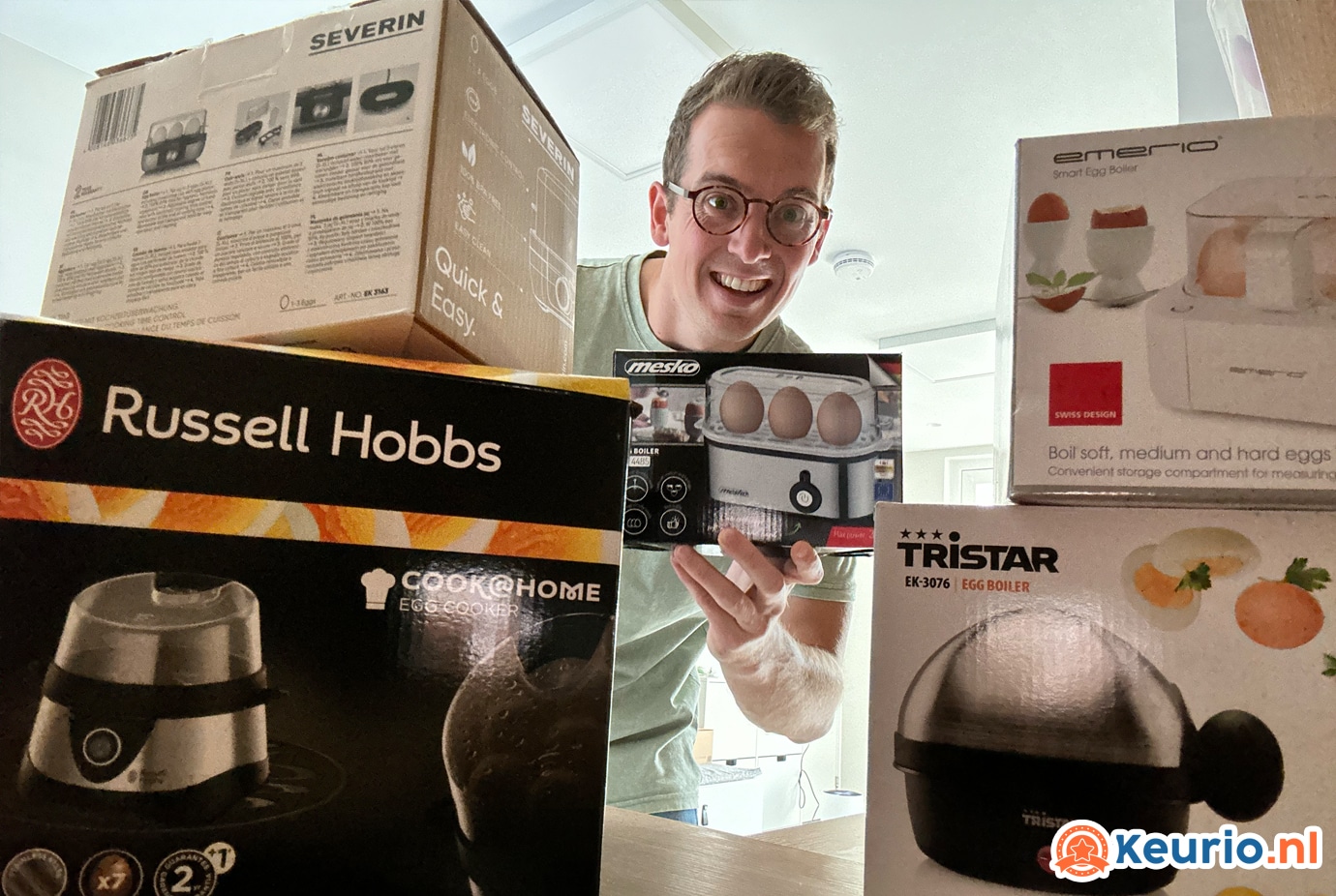 Een man gluurt opgewonden tussen verschillende kleine dozen keukenapparatuur, waaronder merken als Russell Hobbs en Severin, in een goed verlichte kamer.