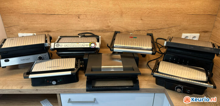 Een verzameling van zes verschillende contactgrillen paninipersen staan netjes gerangschikt op een houten aanrecht. De apparaten variëren in grootte, vorm en knopopstelling. Op de achtergrond is een stopcontact zichtbaar en onder de toonbank bevindt zich een lade.