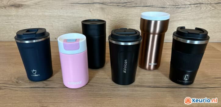Een collectie van zes reismokken, oftewel thermosbekers, van diverse merken en kleuren staan prachtig uitgestald op een houten ondergrond. De mokken hebben ontwerpen in zwart, roze, koper en zwart met merknamen als 'Kambukka', 'Vatten', 'Avocan' en 'Castagna'. Waar laat je op als je er een koopt?