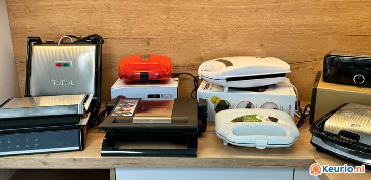 Een opstelling van diverse tafelgrills en tosti-ijzers, waaronder merken als Tefal. Deze apparaten staan op een houten plank, sommige nog in hun dozen. De achtergrond is voorzien van een houten wand en rechtsonder is het logo van "keurio.nl" zichtbaar.