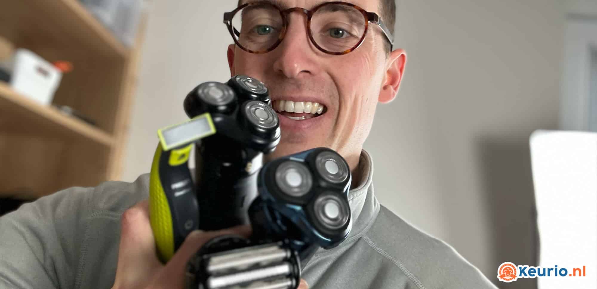Een vrolijke man met bril houdt drie scheerapparaten richting de camera en laat ze met een brede, vrolijke glimlach zien in een huiselijke setting.
