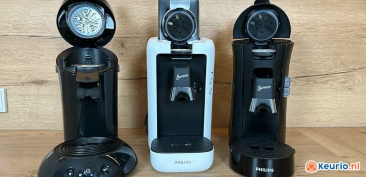 Een zij-aan-zijaanzicht van drie koffiemachines op een houten oppervlak. De machine in het midden is wit met een zwarte voorkant, terwijl de andere twee volledig zwart zijn. Ze dragen allemaal het merk "Philips" met "Senseo" op de voorpanelen. De machine links is open.