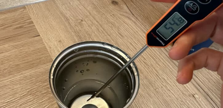 Een hand houdt een digitale thermometer vast, die een temperatuur van 29,4 °C aangeeft, terwijl de metalen sonde is ondergedompeld in een roestvrijstalen beker met vloeistof. De beker wordt op een houten oppervlak geplaatst. De afbeelding heeft rechtsonder een watermerk "keurio.nl".