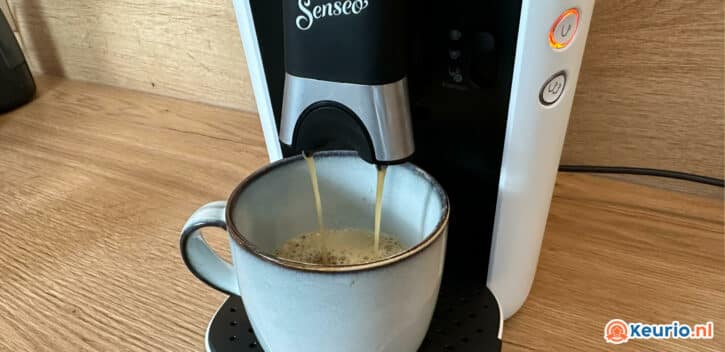 Een koffiezetapparaat met het opschrift "Senseo" schenkt koffie in een keramische kop met handvat. De machine heeft een zwart-wit design en staat op een houten aanrecht. Rechtsonder is een logo van "keurig.nl" zichtbaar.