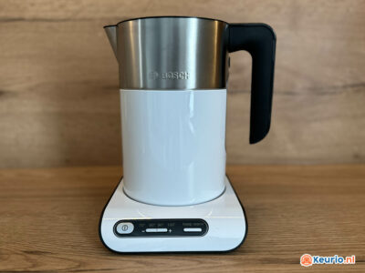 Een witte en zilveren Bosch-waterkoker staat op een houten oppervlak. De waterkoker heeft een zwart handvat en staat op een witte basis met bedieningsknoppen die de temperatuurinstellingen aangeven van 70°C tot 100°C en een "warmhoud" optie. Op de waterkoker is het merklogo zichtbaar.