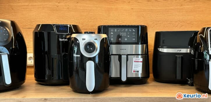 Op een houten plank staat een opstelling van vijf verschillende airfryers tentoongesteld. Elke airfryer heeft een apart ontwerp, variërend in grootte en bedieningselementen, met digitale schermen die verschillende instellingen weergeven. De achtergrond is een houten muur en het logo van Keurio.nl is rechtsonder zichtbaar.