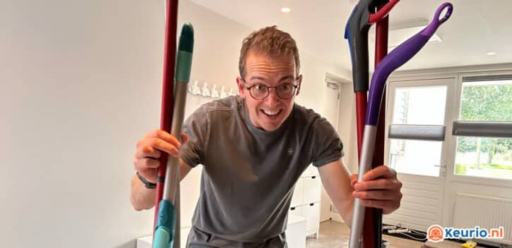 Een persoon met een bril glimlacht terwijl hij verschillende kleurrijke schoonmaakgereedschappen vasthoudt, waaronder wat dweilen. Ze staan in een helder verlichte kamer met witte muren, een raamdeur en wat opbergmeubilair op de achtergrond. Rechtsonder is het logo "keurio.nl" zichtbaar.