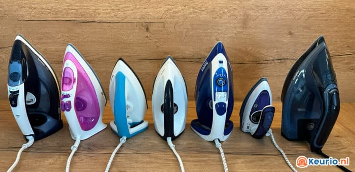 Een rij van zes verschillende elektrische strijkijzers rechtopstaand op een houten oppervlak. Ze variëren in kleuren, waaronder zwart, roze, wit, blauw en grijs. De ijzers zijn uitgelijnd in volgorde van grootte. Rechtsonder is een oranje en wit "keurio.nl"-logo zichtbaar.