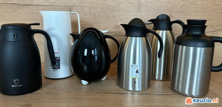 Een rij van vijf thermosflessen op een houten ondergrond. De kolven variëren in kleur en uitvoering: van links naar rechts een zwarte fles met handvat, een witte fles, een zwarte theepotvormige fles en twee roestvrijstalen kolven, elk met zwarte deksels en handvatten.