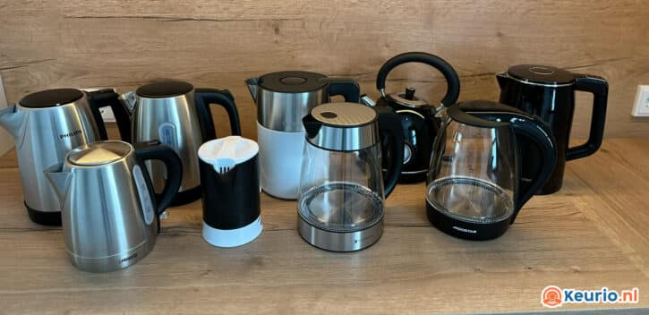Een verzameling van verschillende waterkokers op een houten aanrecht. De ketels hebben verschillende ontwerpen, materialen (roestvrij staal, plastic en glas) en handgreepstijlen. De achtergrond is een houten muur met aan de rechterkant een stopcontact. Rechtsonder is het logo "keurio.nl" zichtbaar.