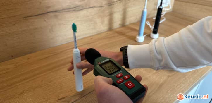 Een persoon houdt een digitale geluidsniveaumeter vast die het geluid van een elektrische tandenborstel meet. De meterstand geeft 29,0 decibel aan. De tandenborstel, wit met een groene opzetborstel, staat op een houten oppervlak en op de achtergrond zijn andere tandenborstels zichtbaar.
