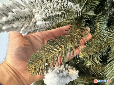 Een close-up van een hand die takken van een kunstmatige kerstboom vasthoudt. Sommige takken zijn bedekt met witte stroompjes die op sneeuw lijken. Op de achtergrond zijn meer groene takken van de boom te zien. Op de afbeelding staat rechtsonder het logo van Keurio.nl.