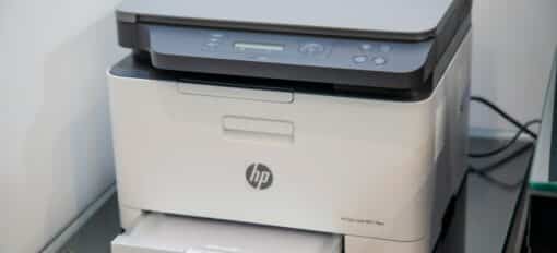 Een moderne HP laserprinter staat op een zwarte tafel tegen een witte muur. Hij heeft een automatische flatbedscanner bovenop, een bedieningspaneel met verschillende knoppen en een klein display, en een papierlade die aan de onderkant iets naar buiten is getrokken.