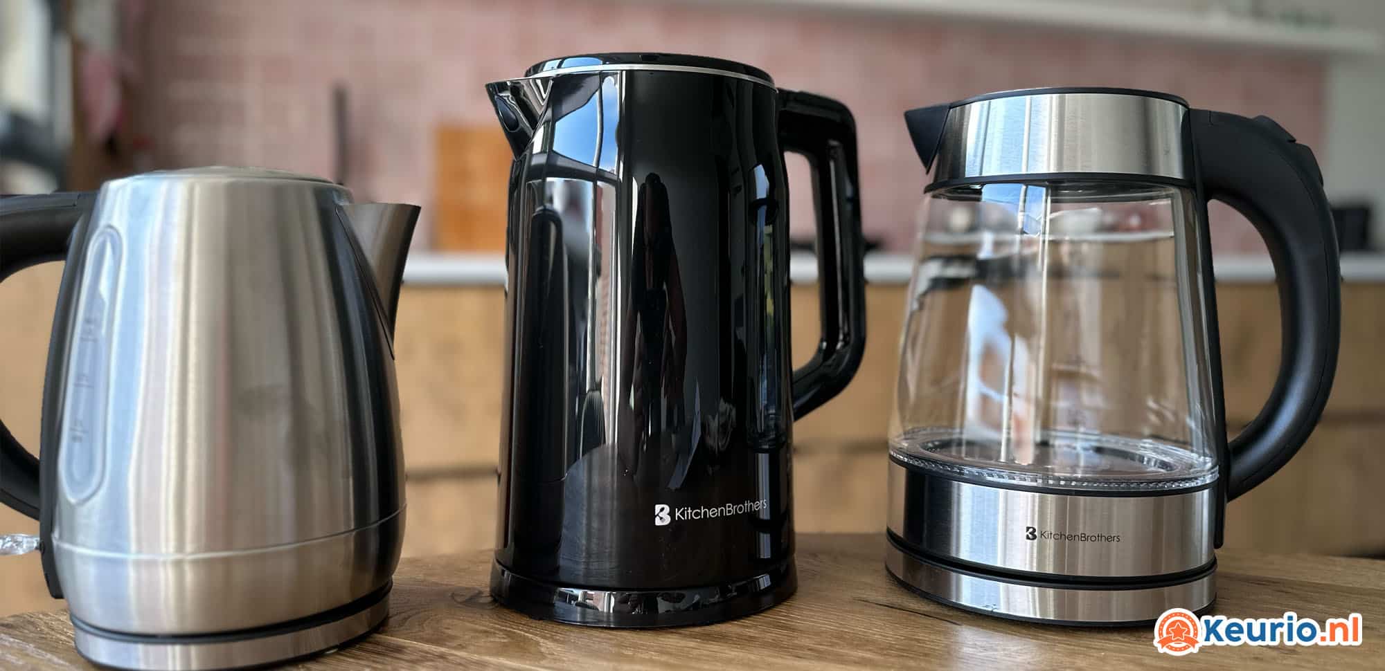 Een close-up van drie verschillende waterkokers op een houten aanrecht in een keuken. De waterkoker-serie bestaat uit één van roestvrij staal, een andere van zwart plastic en de laatste van helder glas en roestvrij staal. Een "Keurio.nl"-logo siert de rechteronderhoek.